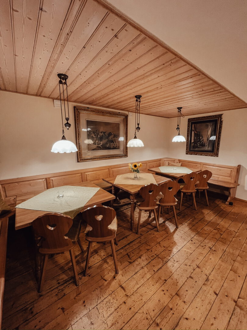3 4er-Tische, jeweils mit einer weißen Tischdecke, darüber eine Hängeleuchte. An der Wand eine Sitzbank, die über die Ecke reicht. Zwei Bilder an der weißen Wand. Helle Holzdielen am Boden und an der Decke.
