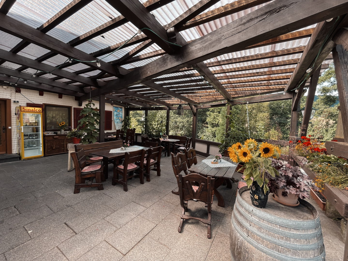 Rustikale Terrasse mit Tischen und Stühlen aus Holz. Überdachung aus Holzbalken. Sonnenblumen im Vordergrund.