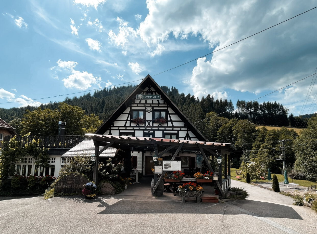 Der Landgasthof "Untere Mühle" von vorne. Ein gemütliches Gasthaus mit Terrasse. Im Hintergrund zeigt sich der Schwarzwald. Blauer Himmel mit einigen weißen Wolken.