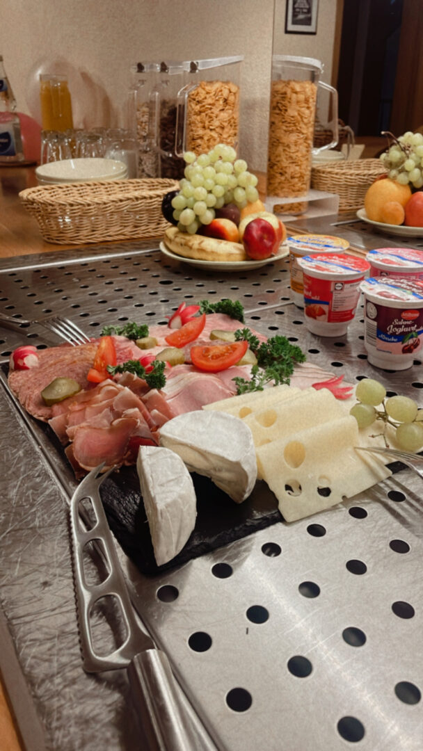 Wurst-Käse-Platte, angerichtet mit Tomaten und Petersilie. Im Hintergrund Obst und Joghurt.