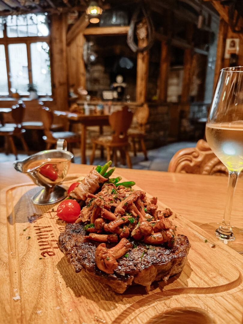 Zwiebelrostbraten mit Bohnen im Speckmantel auf einem Holzbrett. Daneben Weißwein. Im Hintergrund der Blick in die Gaststube.