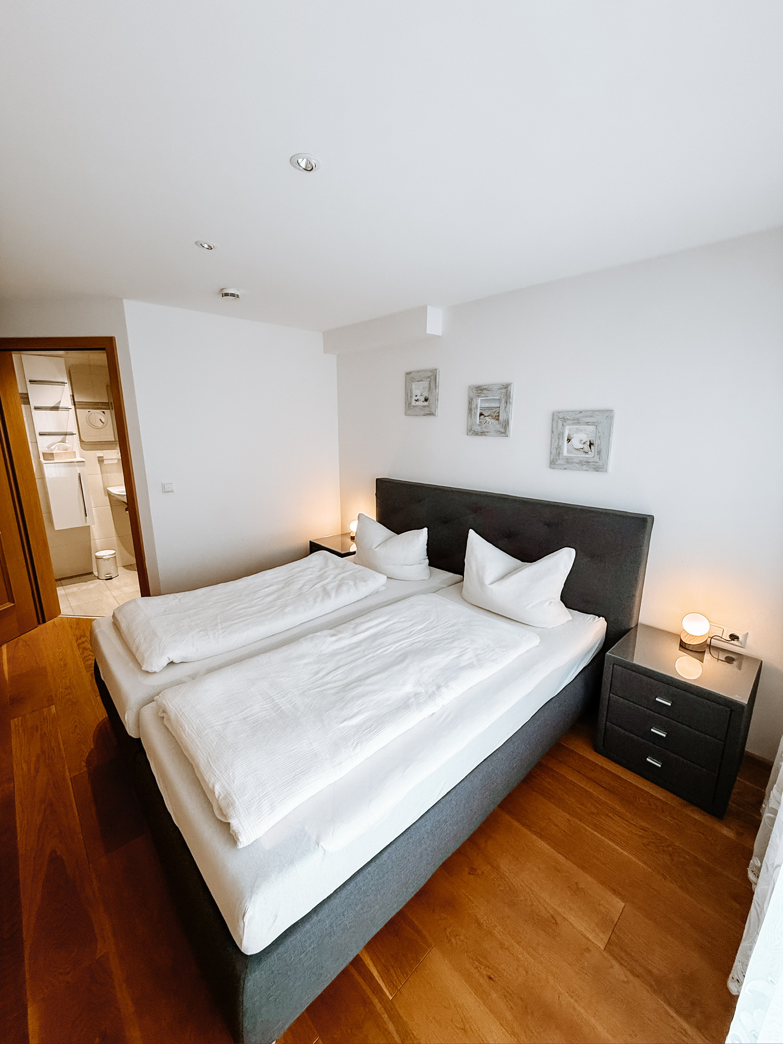 Blick in ein Zimmer mit hellen Holzdielen, einer grauen Couch mit roten Kissen und zwei roten Sesseln. Ein Doppelfenster links und eine Balkontür rechts.