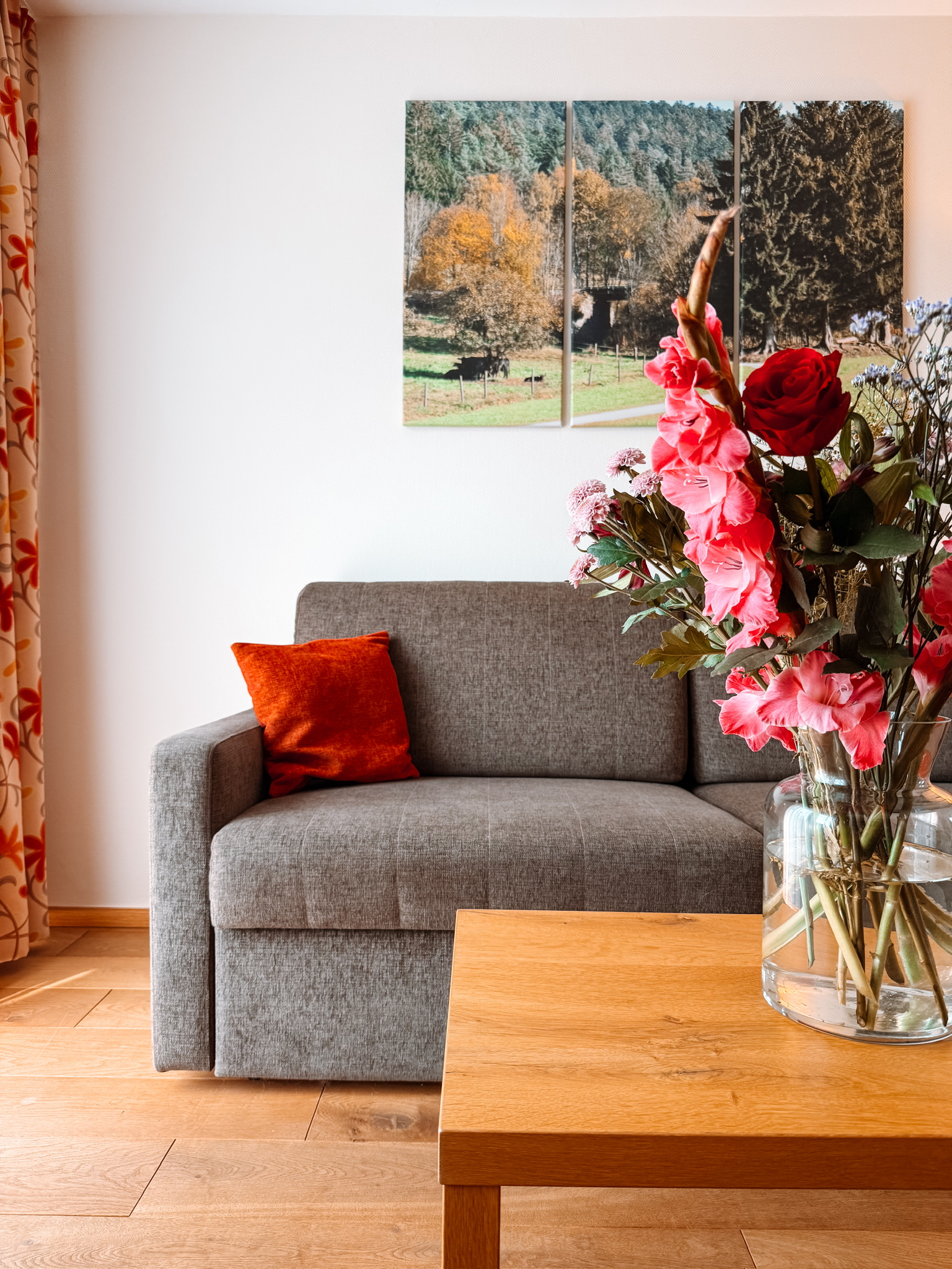 Rosane Blumen in einer durchsichtigen Vase rechts, im Hintergrund ein graues Sofa mit einem roten Kissen. An der weißen Wand hinter dem Sofa hängt ein Bild vom Schwarzwald.