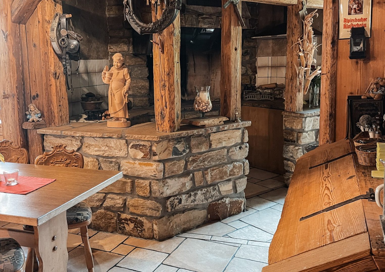 Urige Gaststube mit viel antiker Deko, ein abgetrennter Bereich durch Steine und Holzbalken, dahinter eine alte Küche.