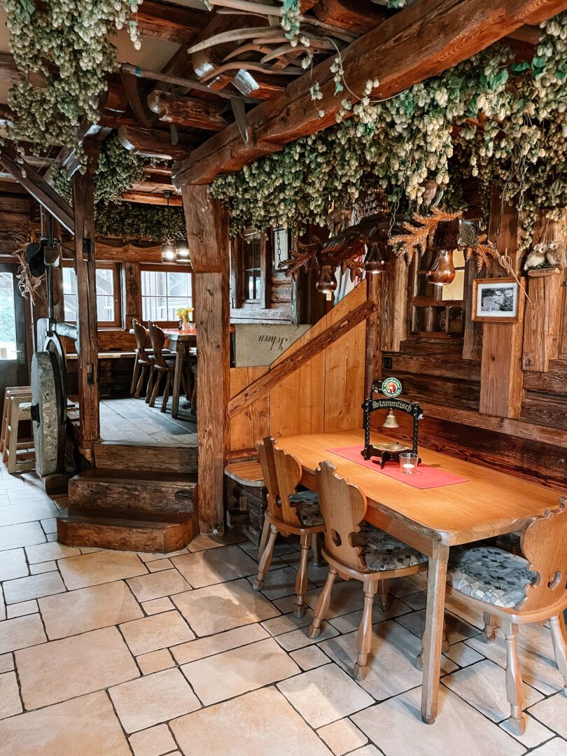 Eine Gaststätte mit einem Holztisch vorne rechts, direkt dahinter eine Holztreppe. Helle Fliesen am Boden. Von oben hängen grüne Äste herunter.