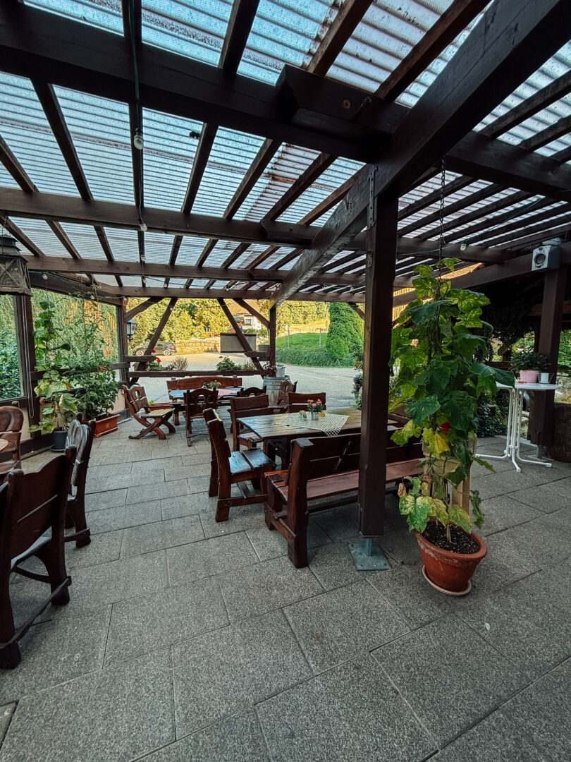 Überdachte Terrasse mit dunklen, robusten Holzgarnituren. Das Dach mit den dunklen Holzbalken wird von senkrechten Balken gestützt.