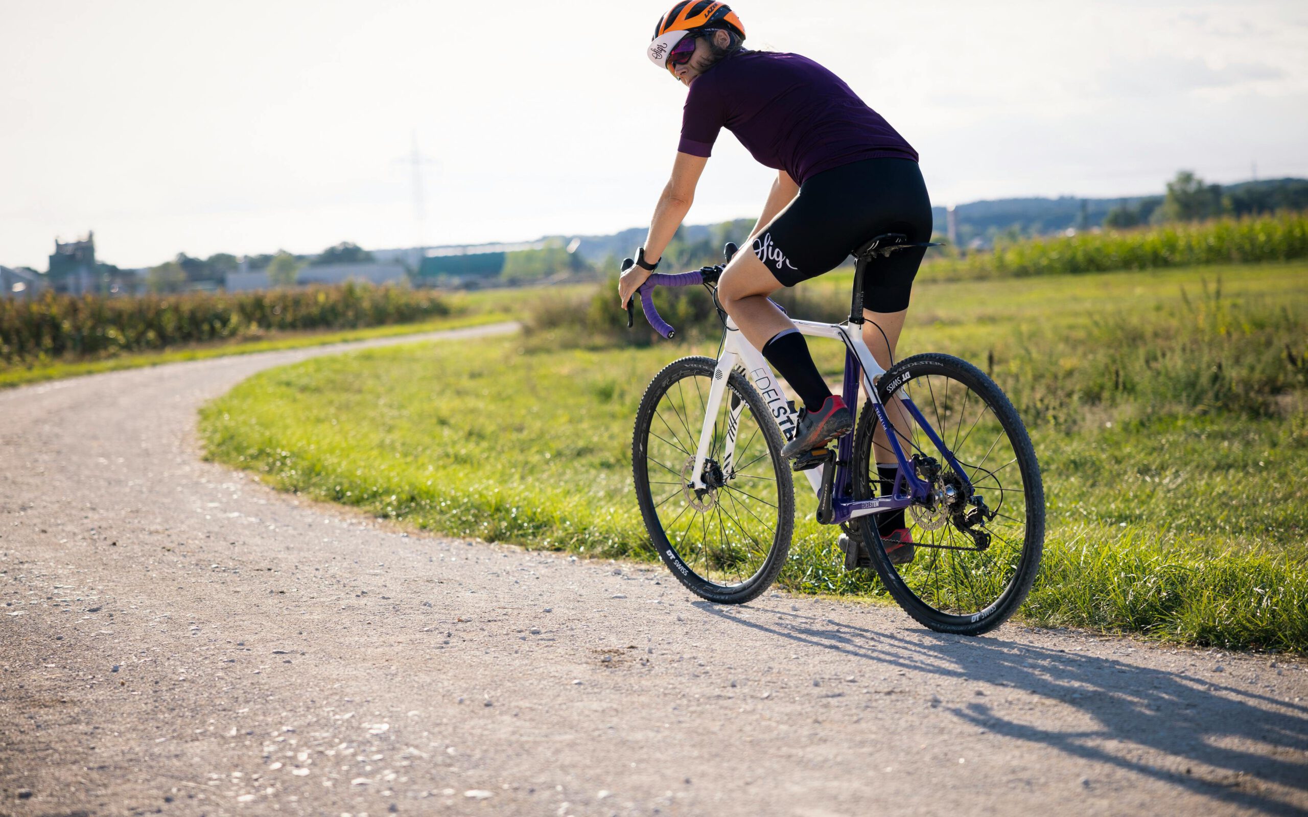 Mann auf einem weißen Fahrrad, sportlich gekleidet mit rotem Helm. Fährt auf einem Schotterweg, daneben eine grüne Wiese.